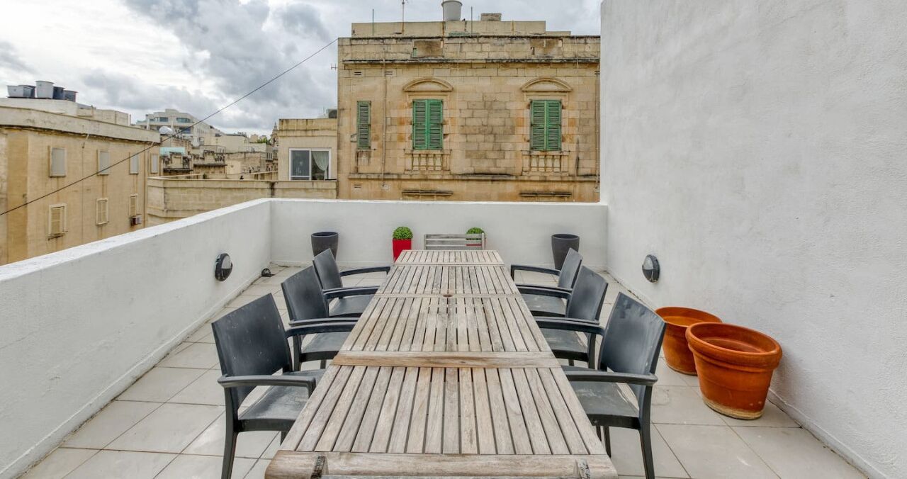 Una terrazza sul tetto con tavolo e sedie in legno, piante in vaso e vista su edifici storici.