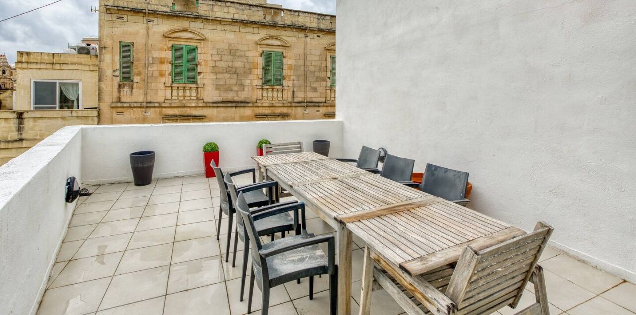 A rooftop terrace with a wooden table and chairs, potted plants, and a view of historic buildings.