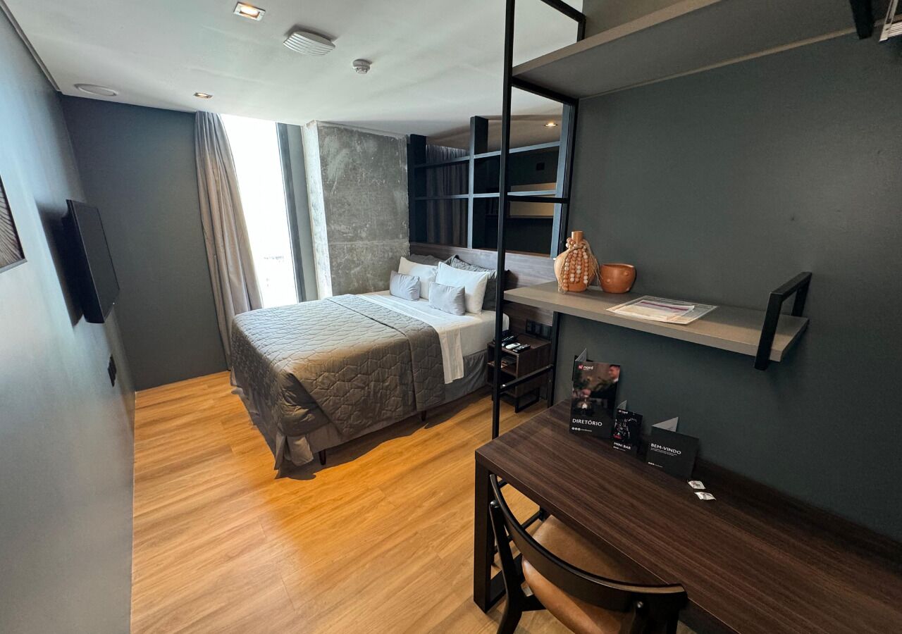 A compact hotel room with a bed, a desk, a chair, a shelf with items, and a window.