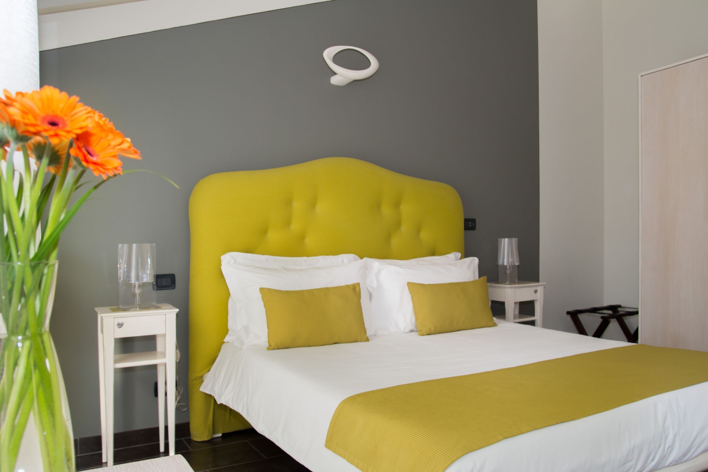A hotel room with a bright yellow headboard, white bedding, and a vase of orange flowers on a bedside table.
