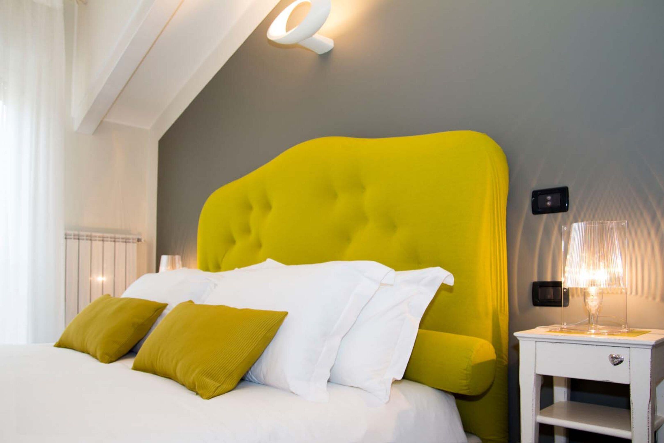 Ein modernes Hotelzimmer mit einem großen Bett, einem leuchtend gelben Kopfteil, weißer Bettwäsche und einem Nachttisch mit einer Lampe.