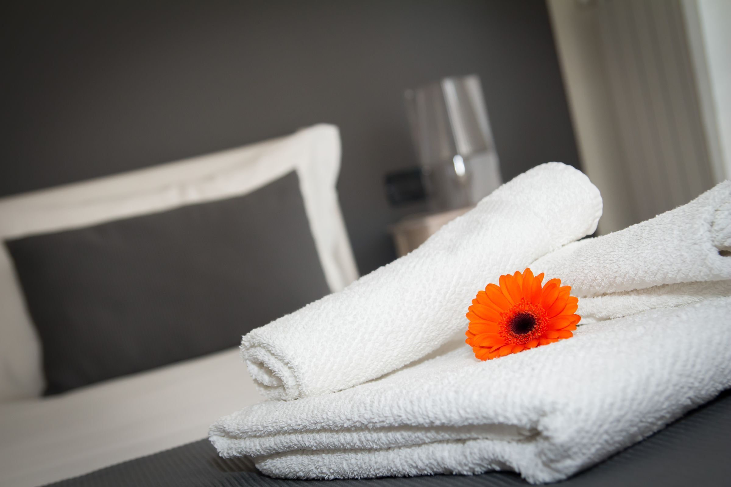 Une pile de serviettes blanches, soigneusement pliées, surmontée d’une seule fleur orange, est posée sur une surface sombre dans une pièce où se trouvent un lit et un oreiller blanc.