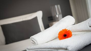 Une pile de serviettes blanches, soigneusement pliées, surmontée d’une seule fleur orange, est posée sur une surface sombre dans une pièce où se trouvent un lit et un oreiller blanc.