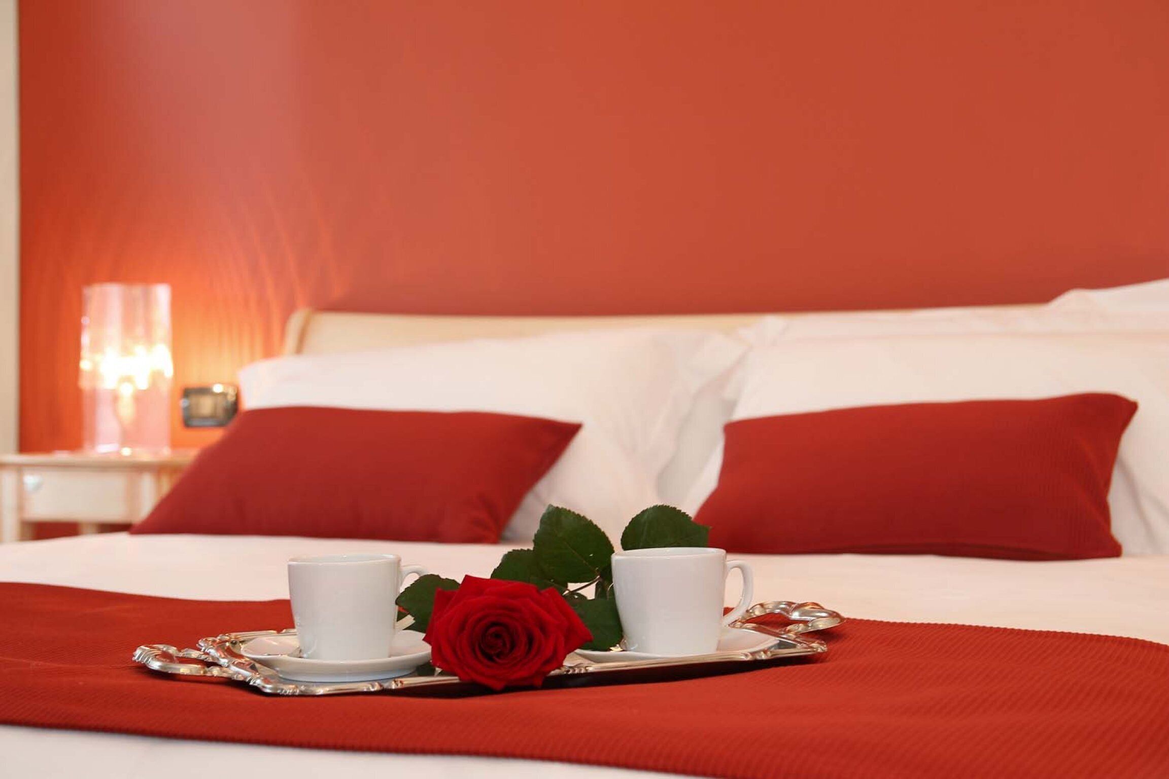 Una cama bien hecha con ropa de cama blanca y roja, dos tazas blancas sobre una bandeja y una rosa roja.