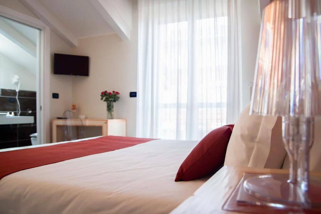 Une chambre d’hôtel avec un lit, un téléviseur fixé au mur, une fenêtre avec des rideaux légers et un vase rempli de fleurs sur une table de chevet.