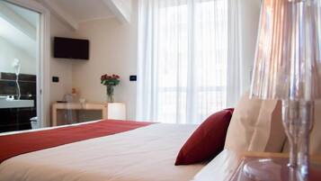 Une chambre d’hôtel avec un lit, un téléviseur fixé au mur, une fenêtre avec des rideaux légers et un vase rempli de fleurs sur une table de chevet.