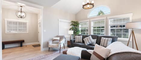 A spacious living room with a grey sofa, a wooden bench, and a large window with blinds.