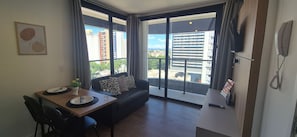 A modern living room with a dining table, black sofa, and large windows offering a city view.