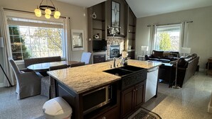 A modern kitchen with granite countertops, a built-in oven, and a large island. There is a dining area with a round table and chairs, and a living room with a sofa and a TV. - Idaho Falls 6bed4bath Exquisite Home (Idaho Falls)