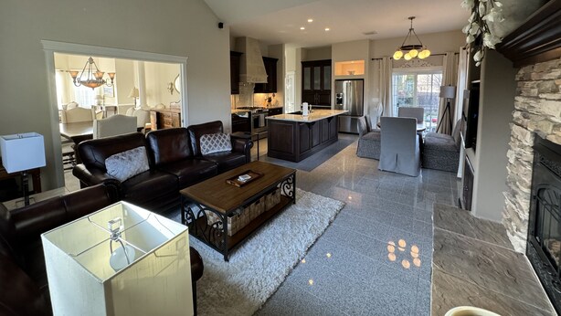 A spacious living room with a dark leather sofa, a wooden coffee table, and a glass-top table. A kitchen with dark cabinets and a central island is visible in the background. - Idaho Falls 6bed4bath Exquisite Home (Idaho Falls)