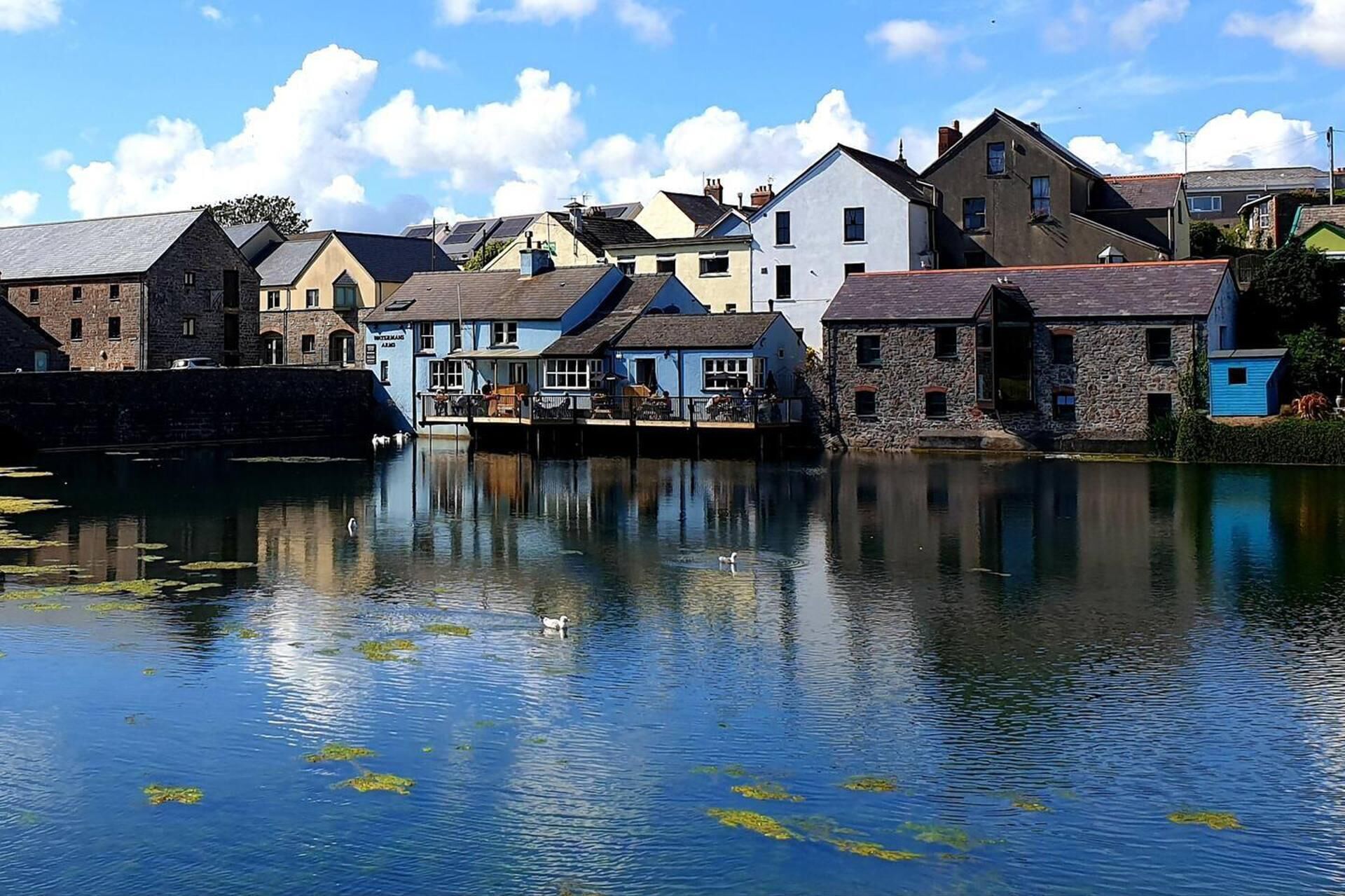 A riverside town with stone buildings, a dock, and calm water reflecting the sky.