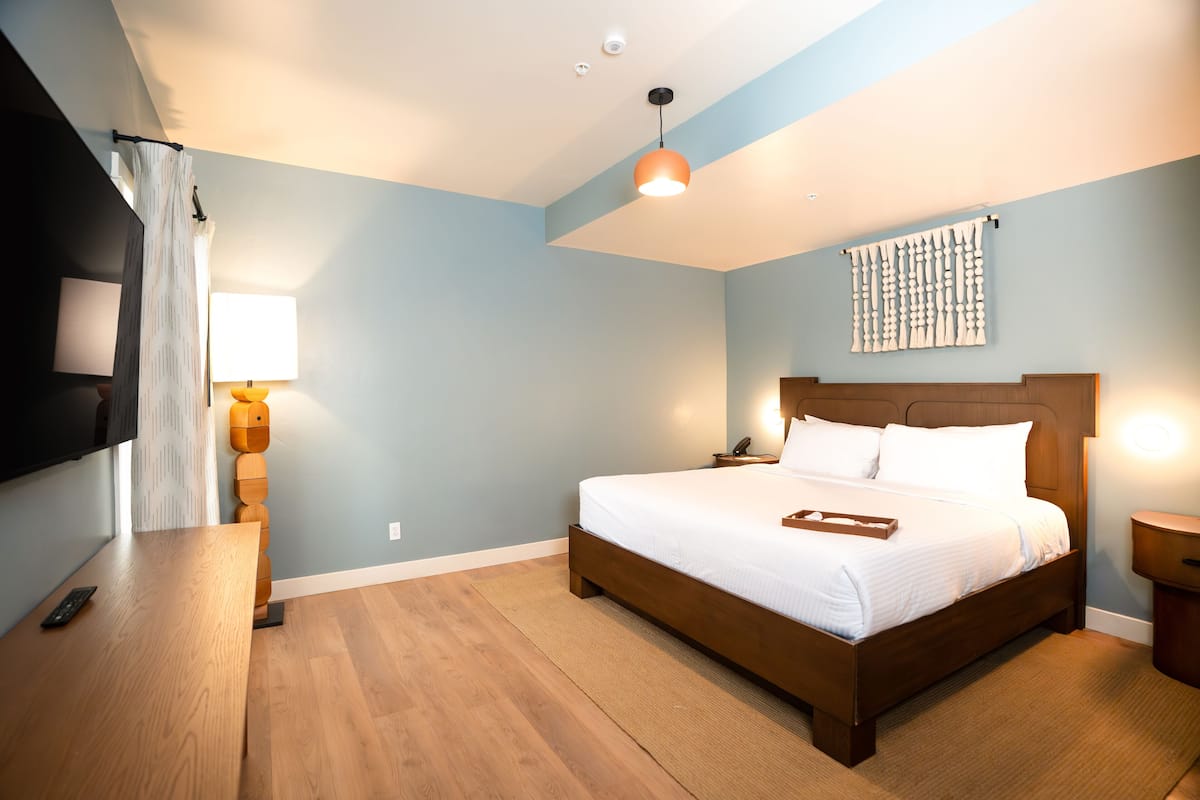 A modern hotel room with a wooden bed, a flat-screen TV, a bedside table, and a wall-mounted artwork.
