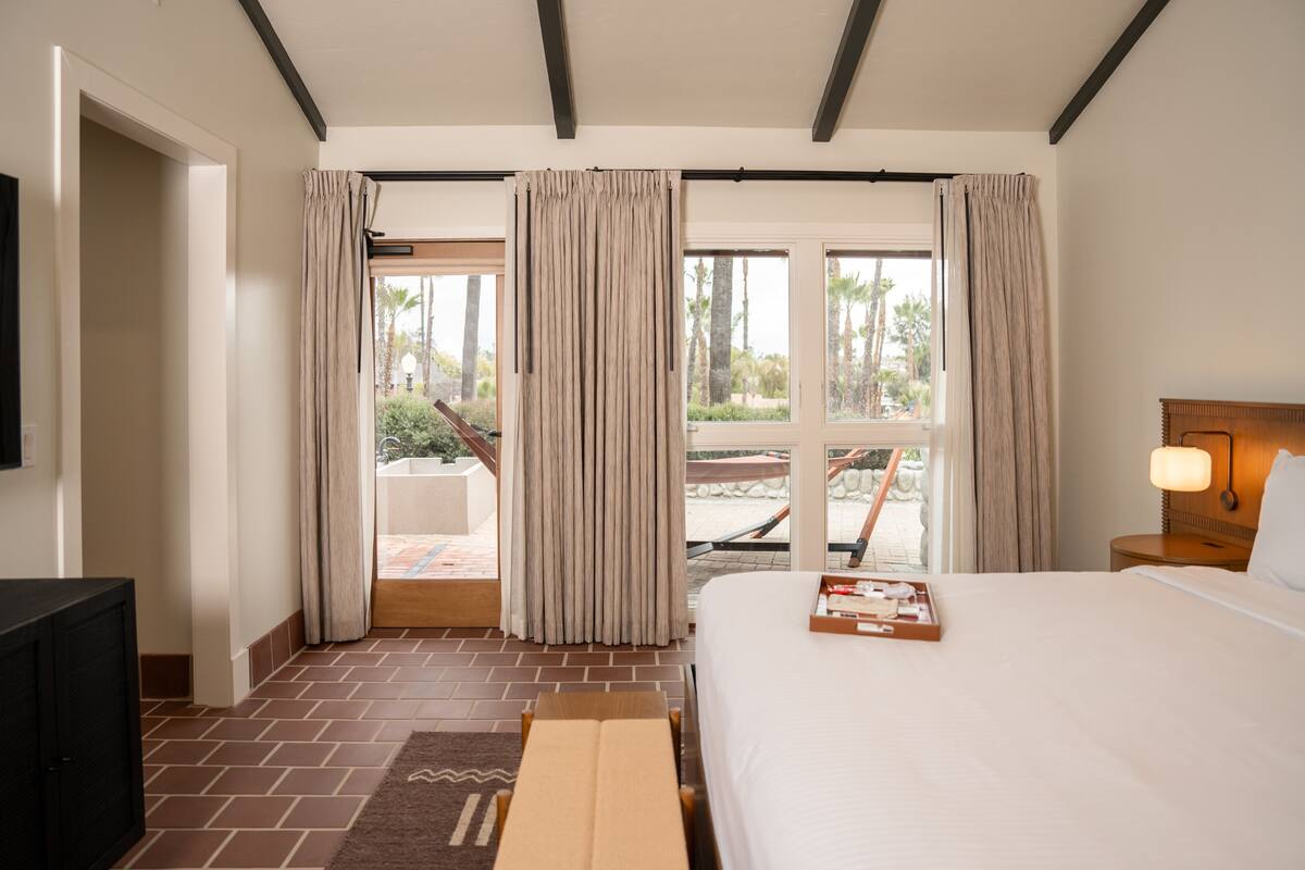 A hotel room with a bed, a TV, a wooden bench, and a balcony with curtains.