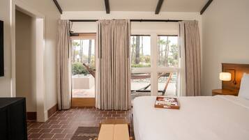 Habitación de hotel con cama, televisión, banca de madera y balcón con cortinas.