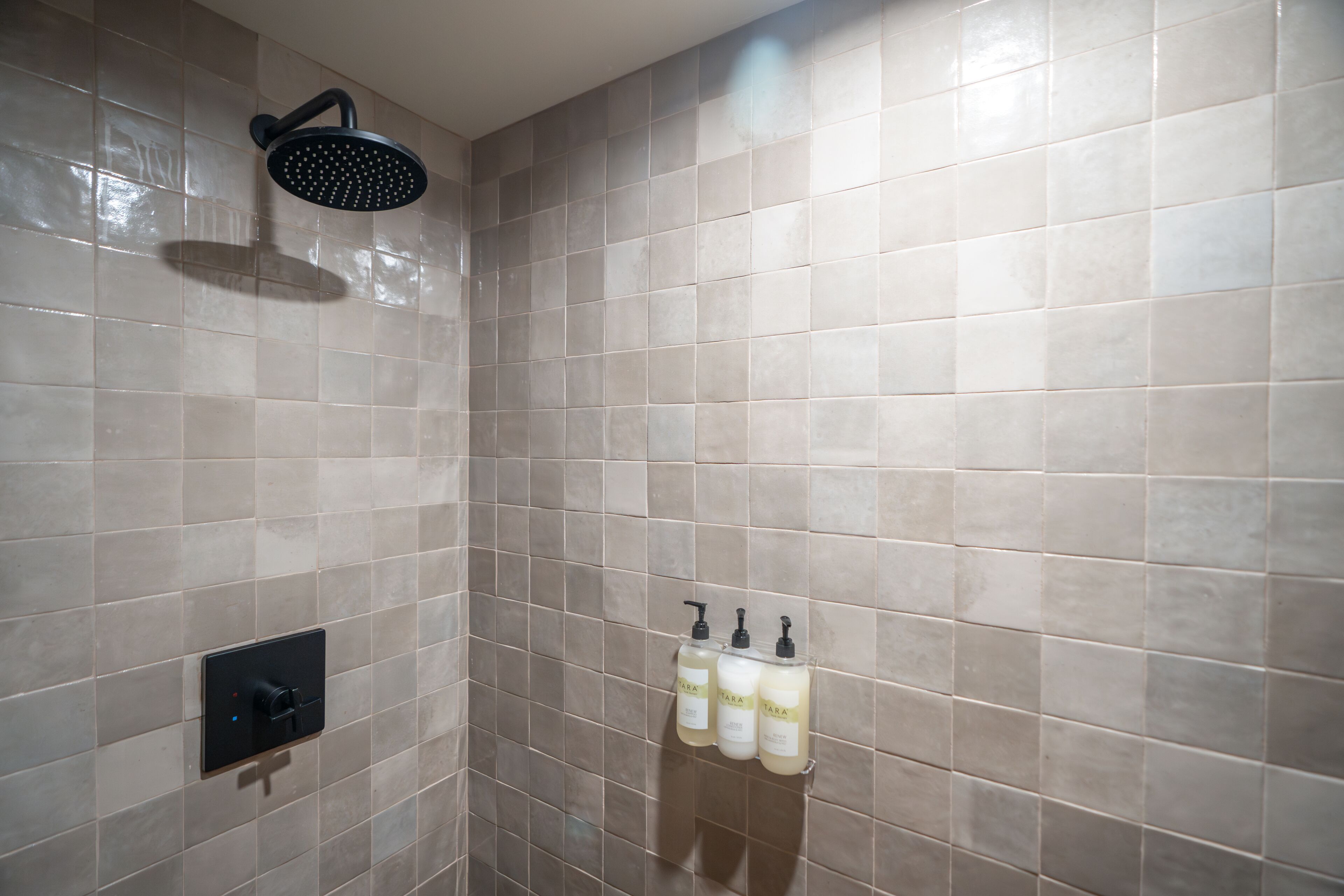 A bathroom with beige tiles, a showerhead, and three bottles of shampoo and conditioner on a wall-mounted rack.
