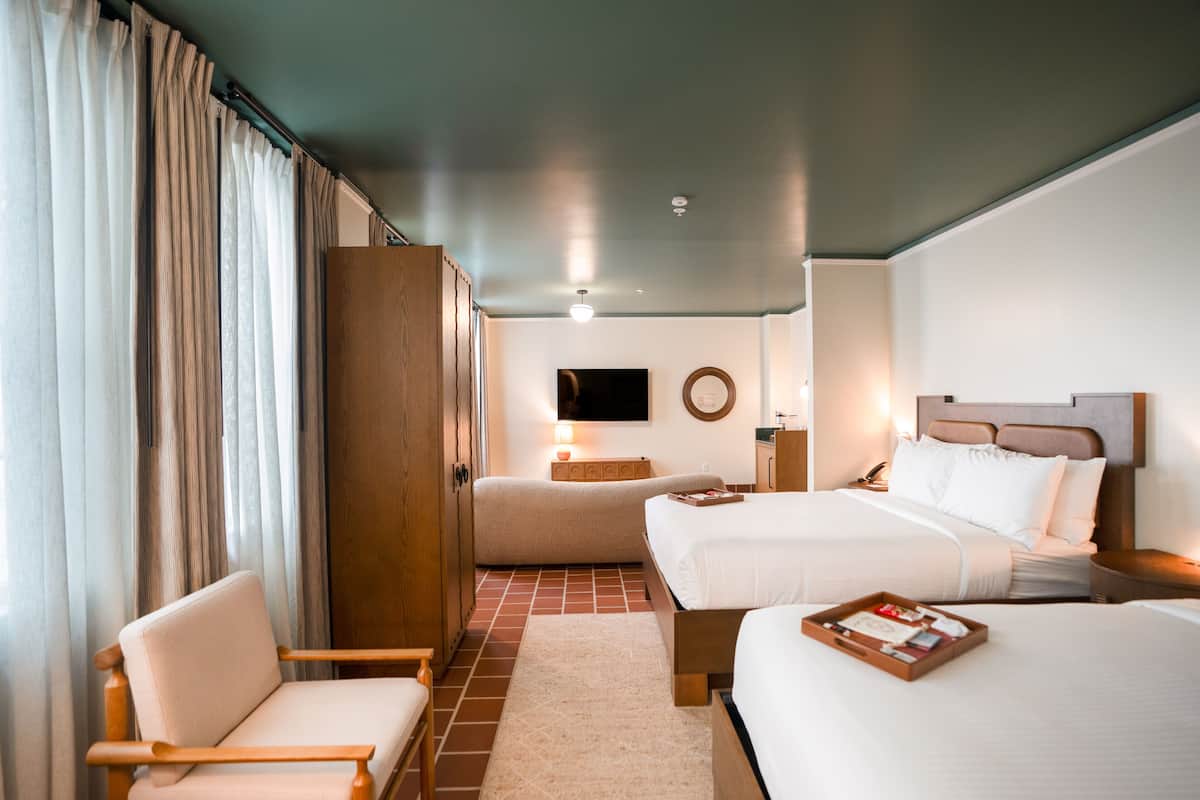 A hotel room with two beds, a wooden wardrobe, a chair, a TV on the wall, and a bedside table with a lamp.