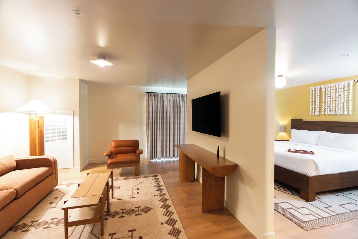 A modern hotel room with a sofa, a wooden bench, a TV mounted on the wall, and a bed with a headboard.