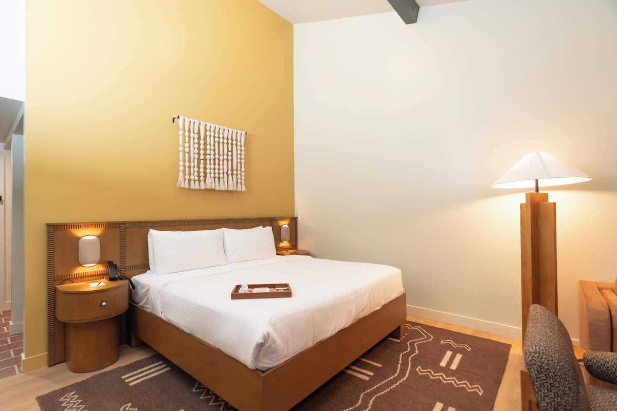 A neatly made bed with white linens, a bedside table with a lamp, and a patterned rug on the floor.