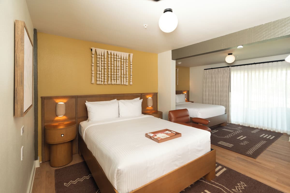A hotel room with a large bed, two bedside tables with lamps, a headboard with a decorative panel, a brown rug, and a window with curtains.