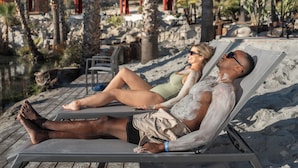 Una pareja descansando en una silla de sol junto a la piscina, con palmeras y vistas a una zona turística.