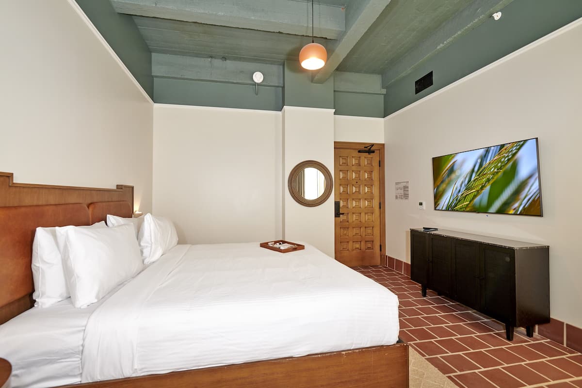 A neatly made bed with white linens, a wooden headboard, a bedside table, a wall-mounted TV, a mirror, and a door with a grid pattern.