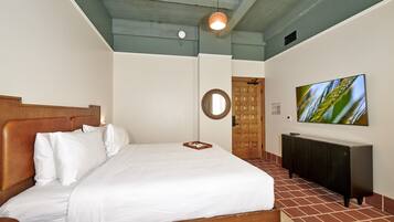 Un lit bien fait, avec du linge de lit blanc, une tête de lit en bois, une table de chevet, un téléviseur fixé au mur, un miroir et une porte à motifs géométriques.