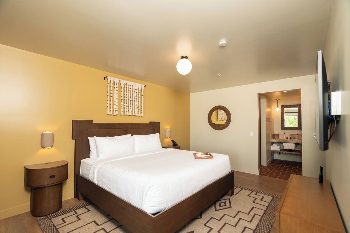 A hotel room with a large bed, bedside lamps, a wall-mounted TV, and a bathroom visible through an open door.