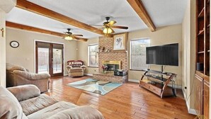 A spacious living room with hardwood flooring, a fireplace, a large flat-screen TV, and a ceiling fan. - New! Spacious 3K sq ft 4BD/3Bath + Pool (Bakersfield)