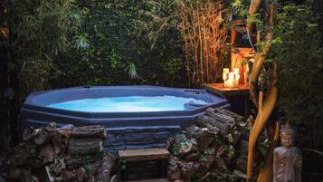 A hot tub surrounded by logs and greenery, with a statue and candles on a wooden table nearby.
