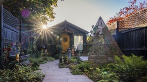 A garden pathway leading to a wooden house with a yellow door, surrounded by lush greenery and a decorative metal structure. - Romantic Lodge on Edge of Nature Reserve (Colchester)