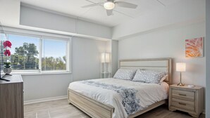 A bedroom with a bed, bedside tables, a ceiling fan, a window with blinds, and a view of trees. - Totally Renovated 1BR/1BA/All New Furniture/Sofa Bed/Balcony/Walk-In Shower/WiFi (Hilton Head Island)