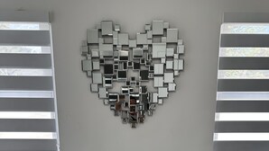Un área de comedor moderna con una mesa de vidrio y sillas de metal, un espejo con forma de corazón en la pared y ventanas con persianas.