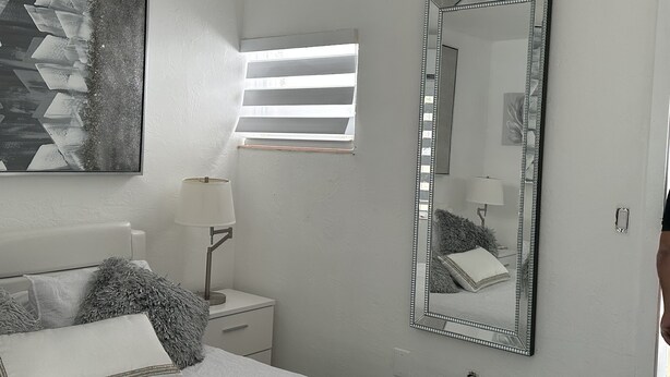 Un dormitorio con cama, mesita de noche, lámpara, espejo grande y ventana con persianas.