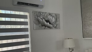 Una habitación con ventana con persianas horizontales, aire acondicionado montado en la pared, un cuadro blanco y negro de flores enmarcado, una mesita de noche blanca con lámpara y una silla blanca.