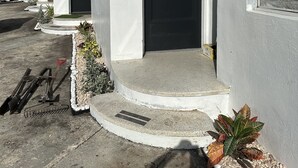 Una hilera de edificios blancos con números en las puertas, un camino de hormigón y un pequeño jardín con plantas y una pala.