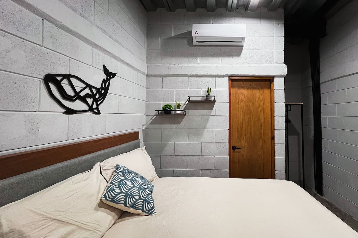 A bedroom with a bed, a wooden door, a wall-mounted air conditioner, and a decorative wall piece.