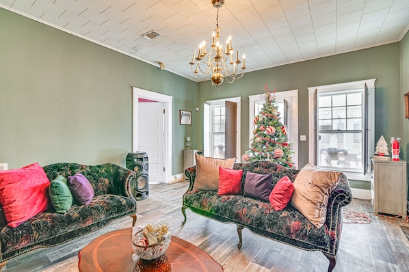 Una sala de estar con dos sofás verdes tapizados, cojines de colores, una araña de techo, un árbol de Navidad y un ventanal con vistas al exterior.