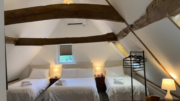 Ein gemütliches Schlafzimmer unter dem Dach mit zwei Betten, Holzbalken, einem Hängesessel und einem Fenster mit Jalousien.