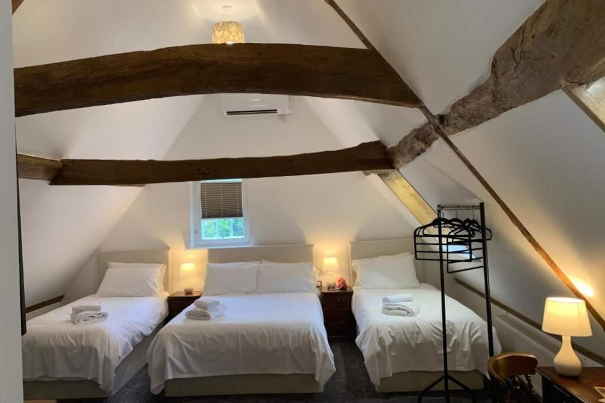 A cozy attic bedroom with two beds, wooden beams, a hanging chair, and a window with blinds.