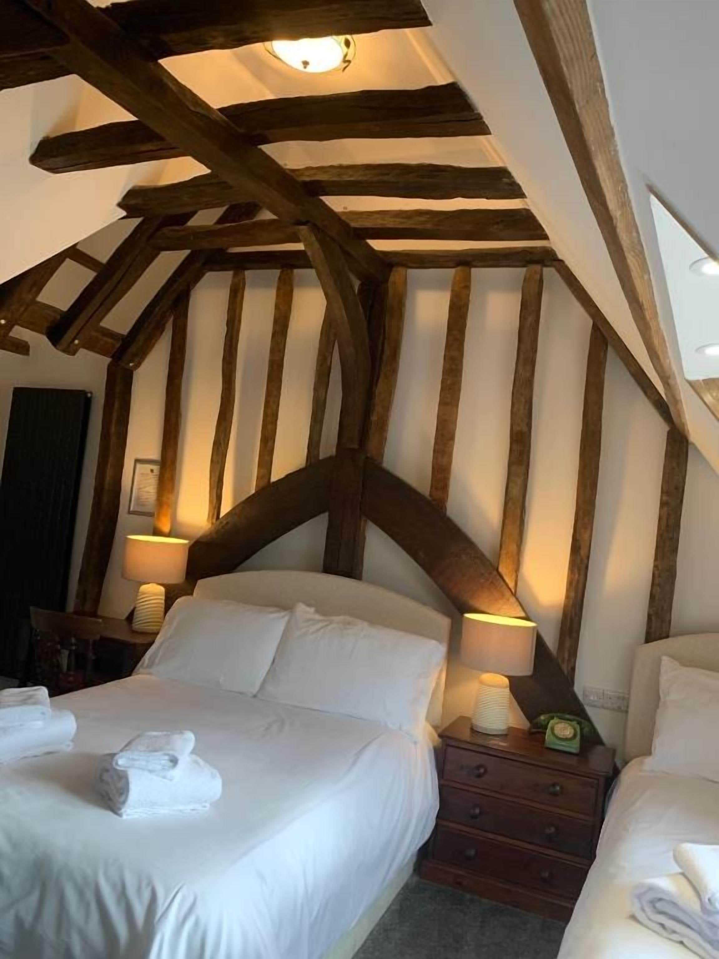 A bedroom with a sloped ceiling, wooden beams, two beds with white linens, bedside tables with lamps, and a nightstand with a clock.