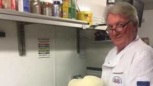 Una persona con bata de chef sostiene una masa grande en una cocina con un mostrador cubierto de harina y varios recipientes en estanterías.