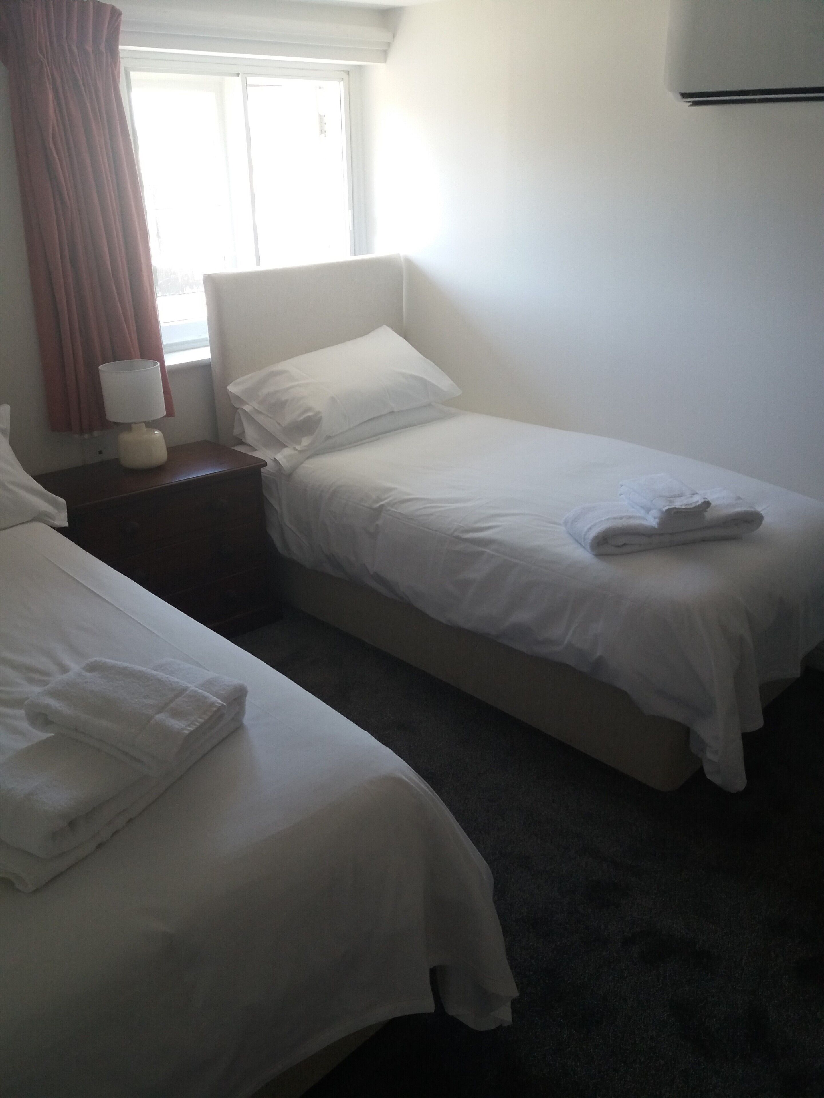 A twin bed with white linens and a folded towel, a bedside table with a lamp, and a window with curtains.