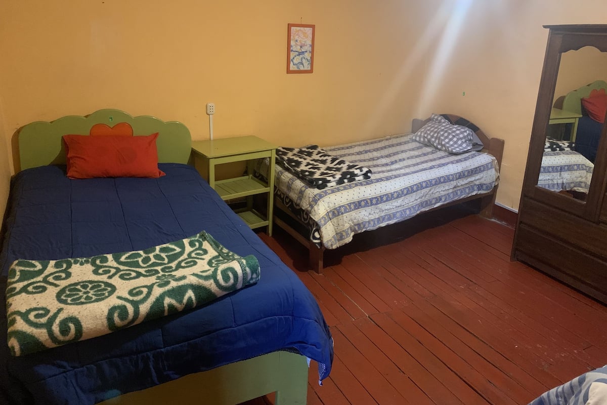 A room with two beds, one with a green headboard and the other with a wooden headboard. There is a small table with a green top and a wall-mounted light fixture.