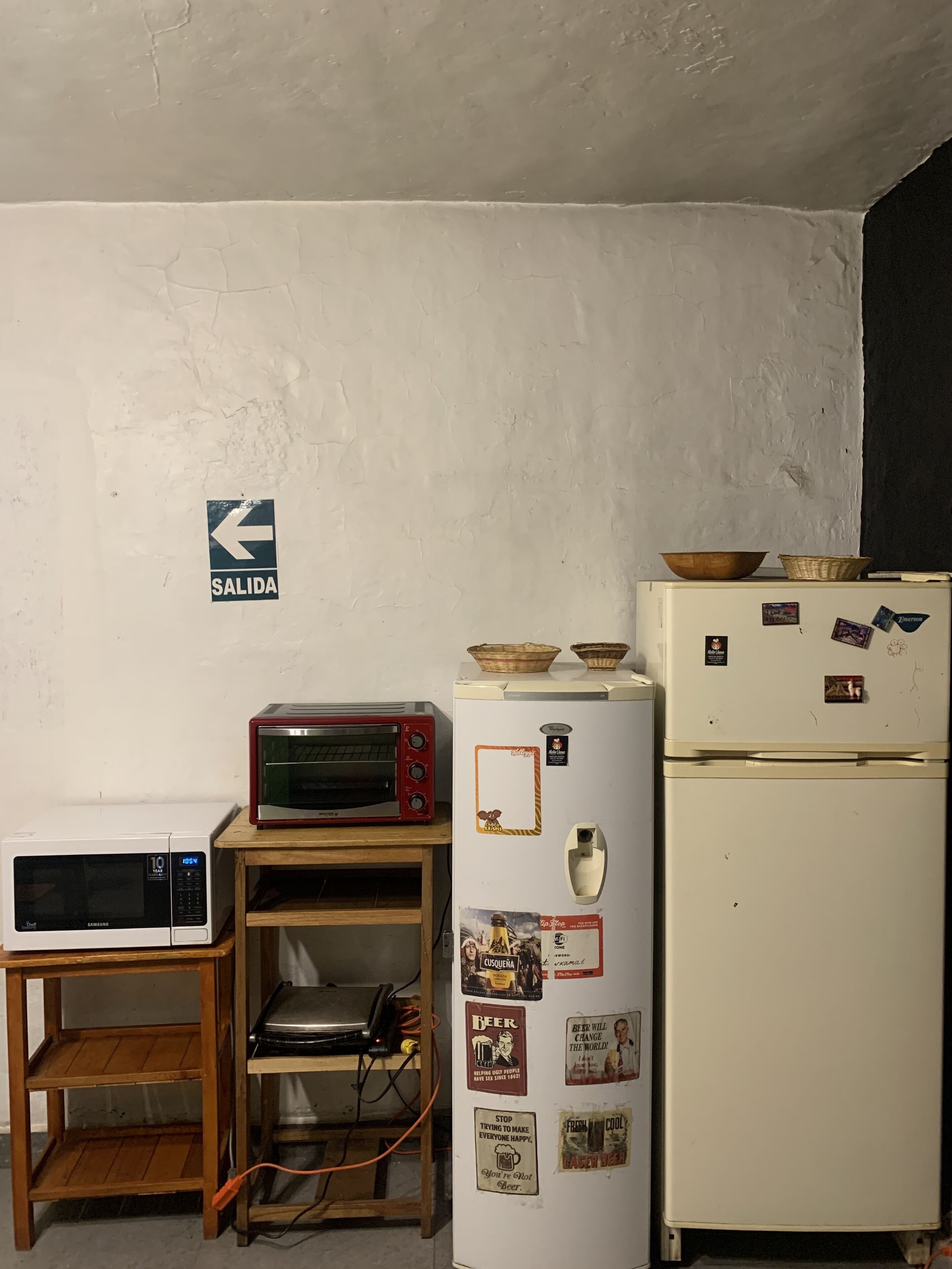 Une petite cuisine équipée d’un four à micro-ondes, d’un grille-pain et d’un réfrigérateur, une étagère en bois et un panneau indiquant la « SORTIE » sur le mur.