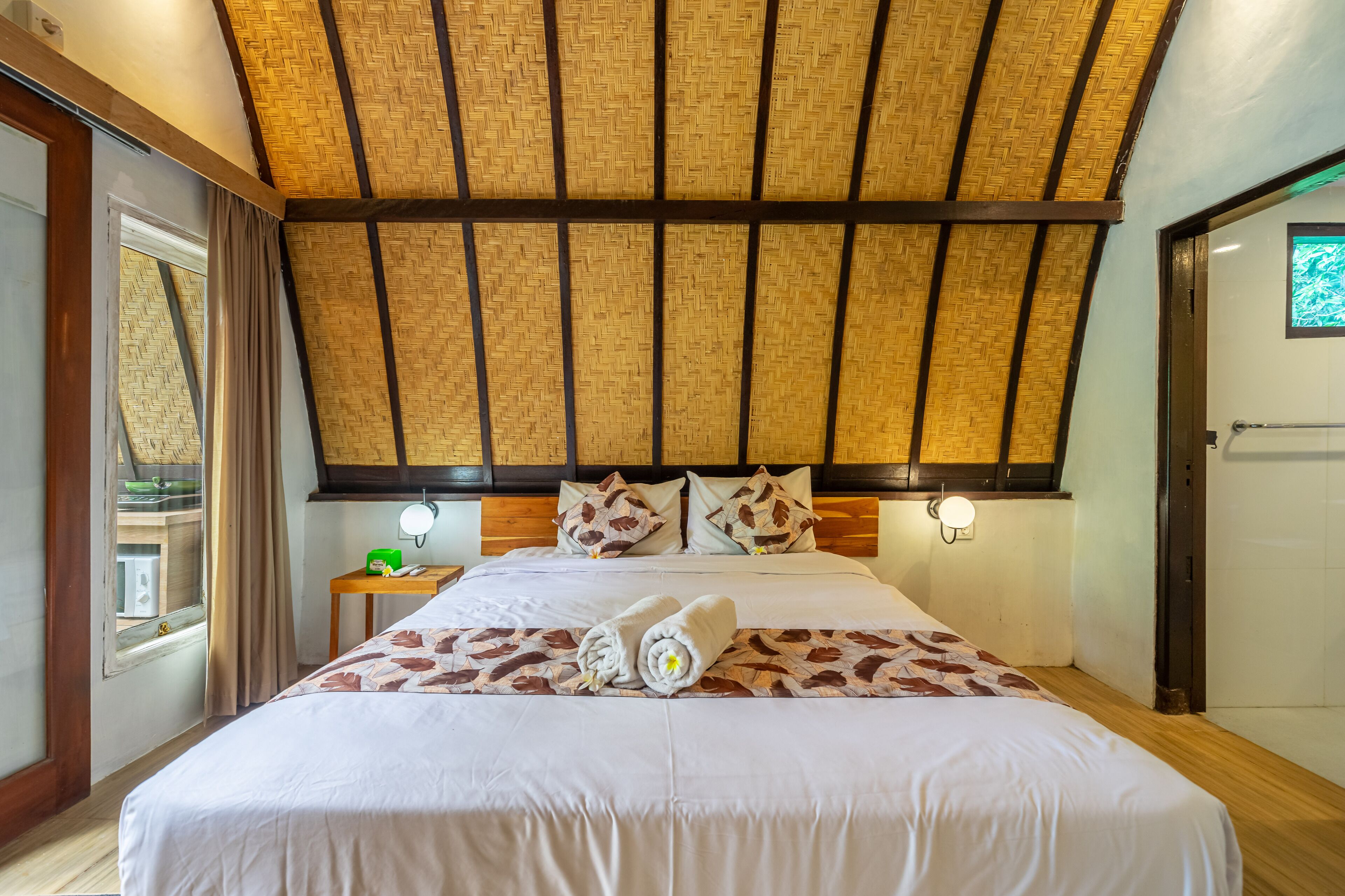 A hotel room with a large bed, a wooden ceiling, a nightstand with a lamp, and a window with curtains.