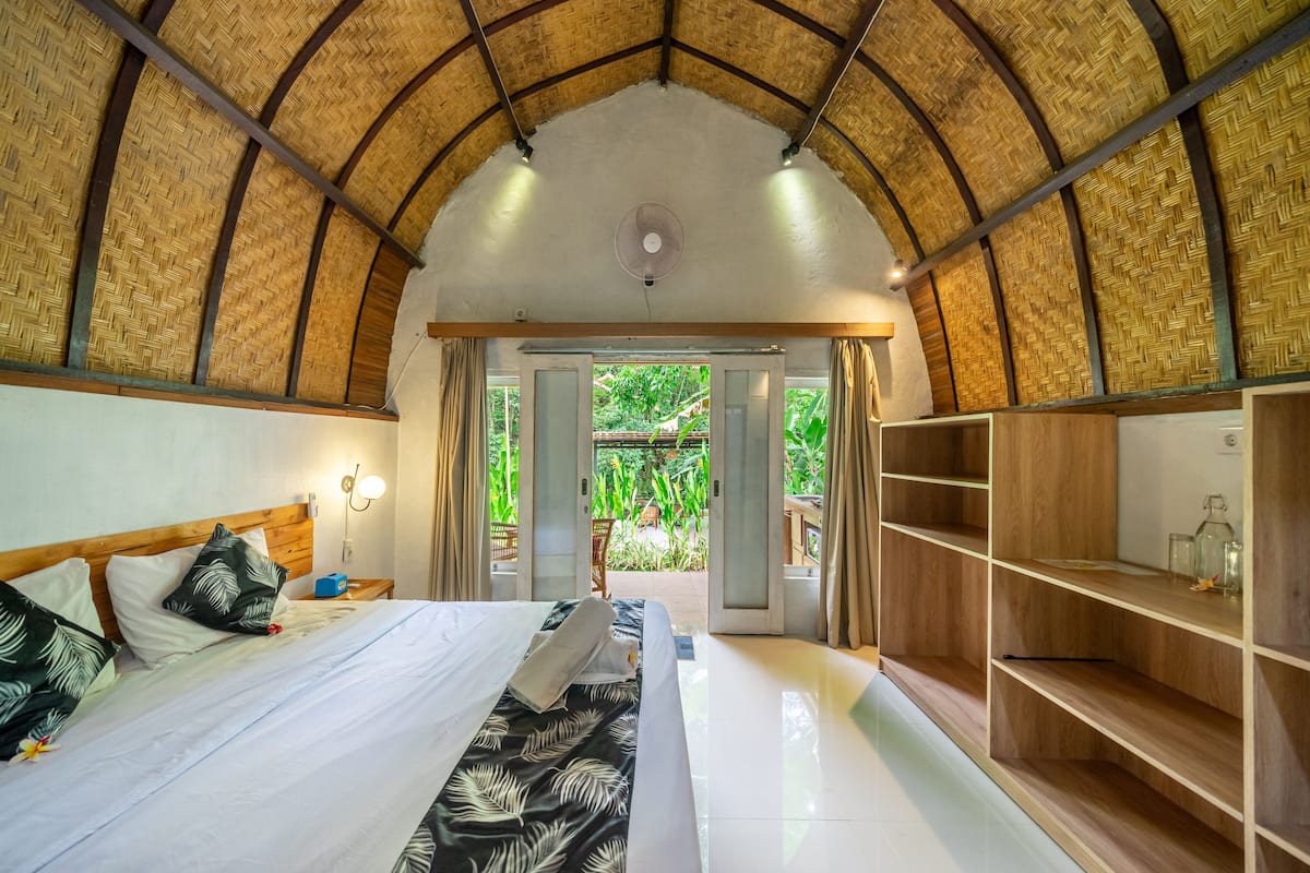 A room with a high, woven ceiling, a bed with white linens, a wooden shelf, and a view of greenery outside.