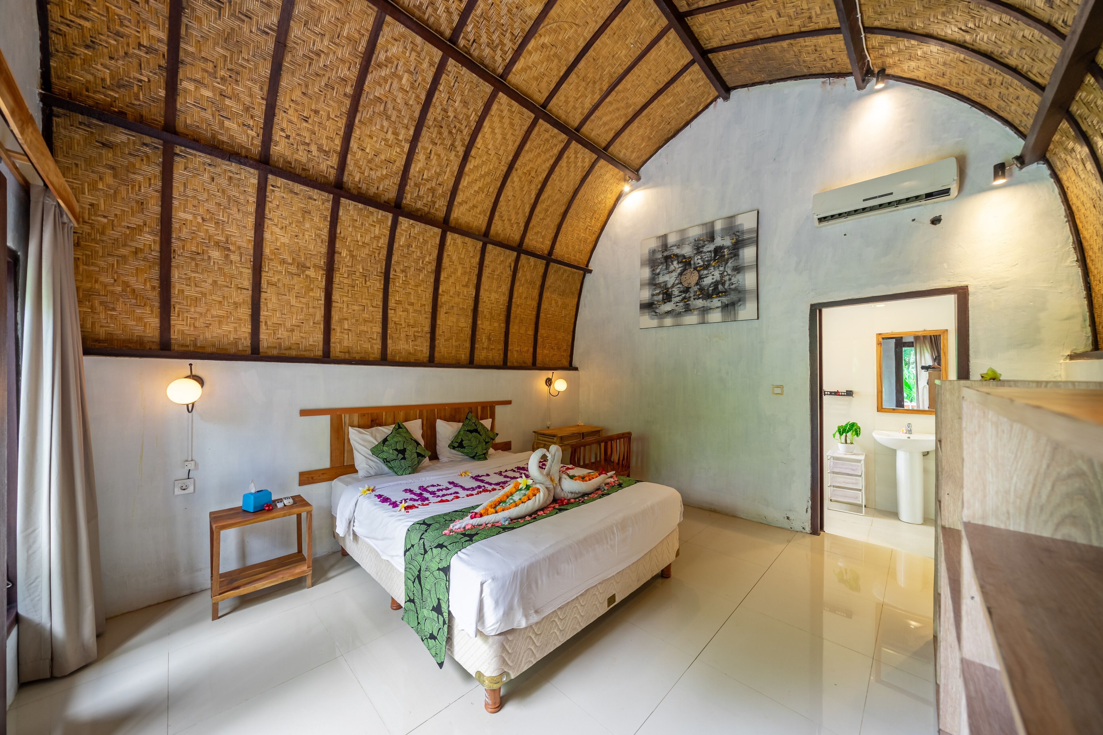 A bedroom with a bed, bedside tables, a nightstand, a mirror, a sink, and a shower area.