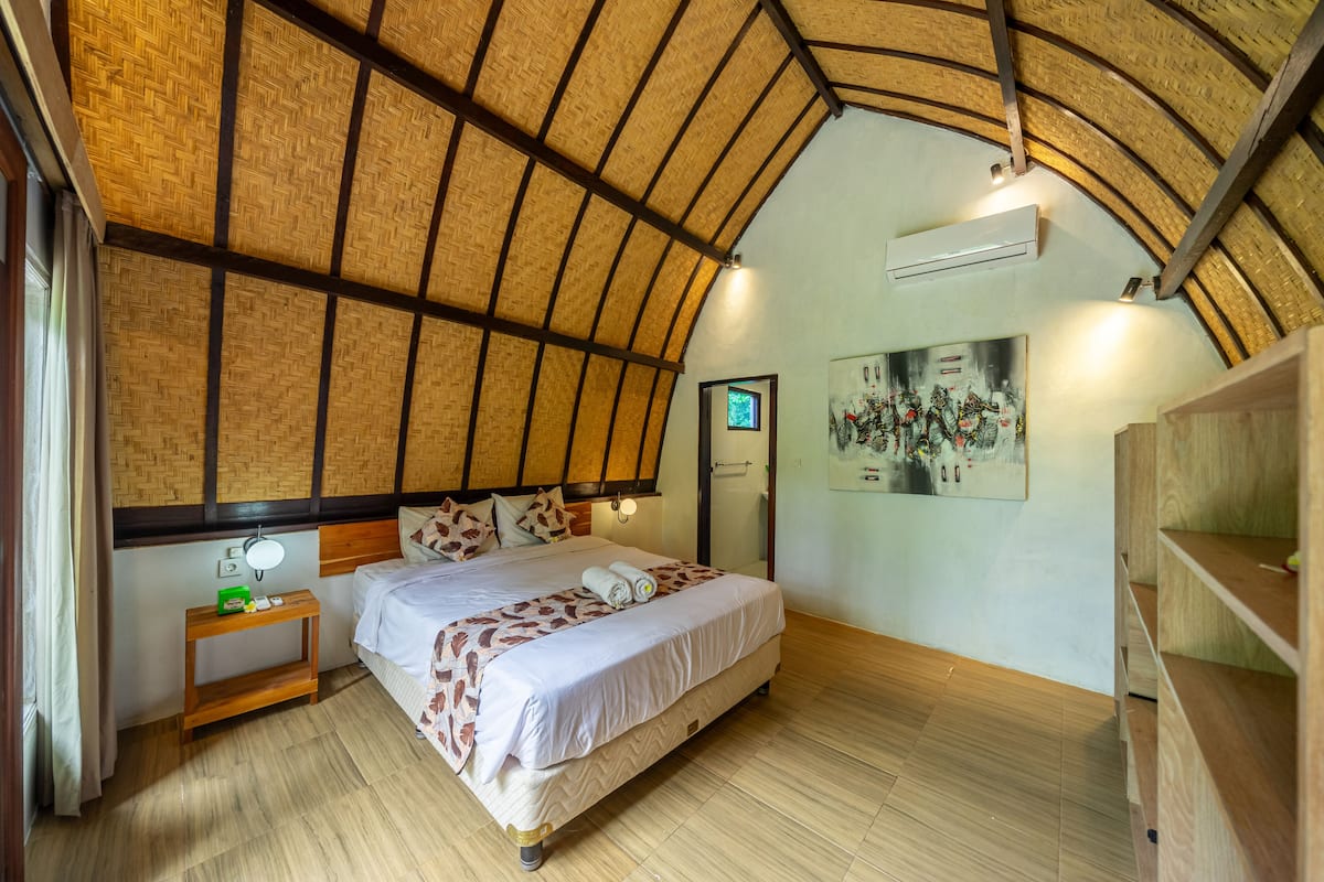A bedroom with a high, curved ceiling, a bed with a patterned bedspread, a bedside table with a lamp, and a wall-mounted TV.