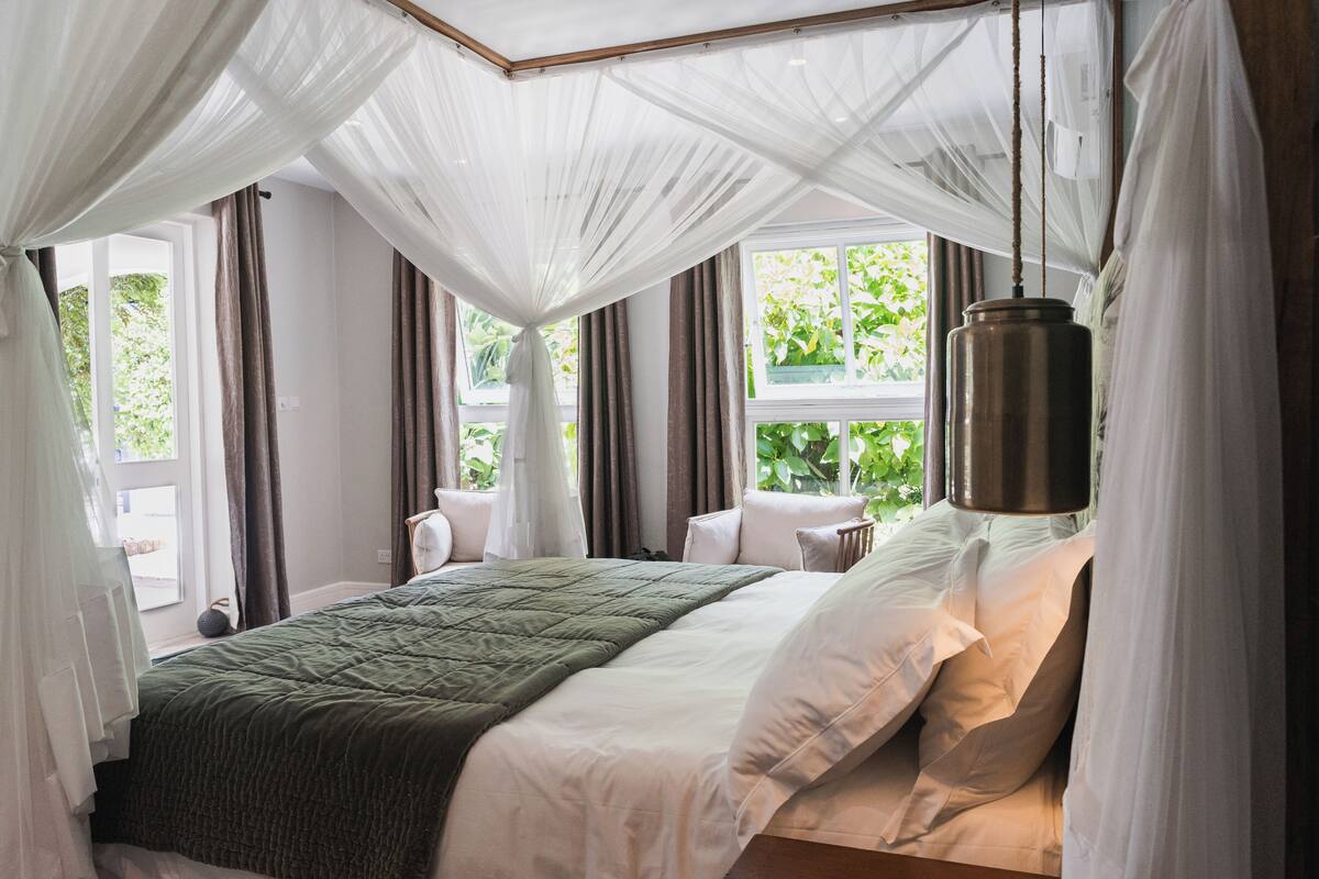 A bedroom with a bed under a canopy, a hanging lamp, and a view of greenery outside.