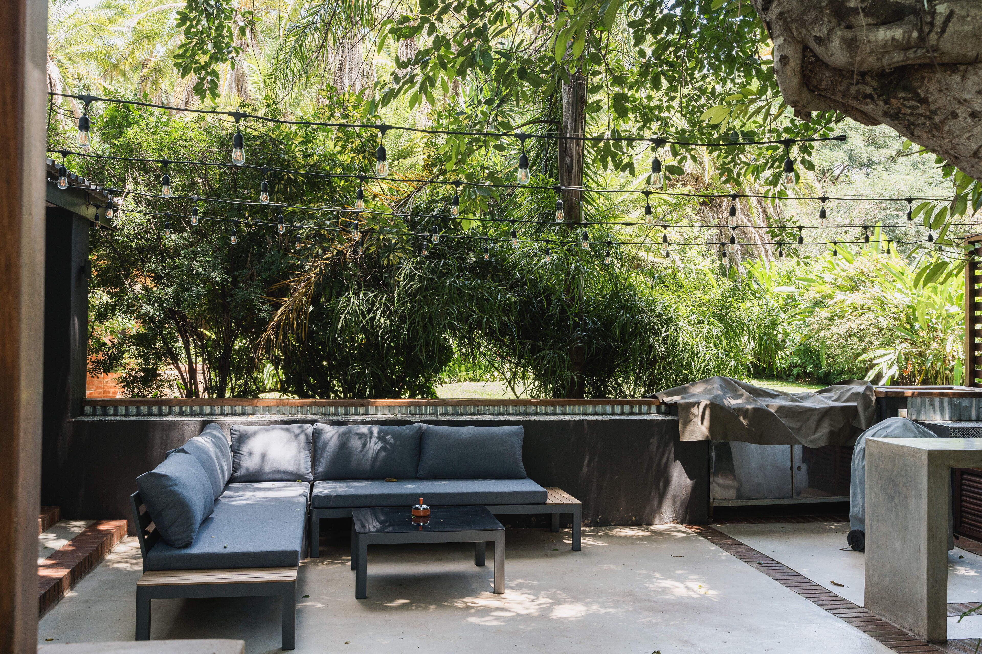 Un espacio al aire libre moderno con sillones y una mesa, rodeado de abundante vegetación y luces colgantes.